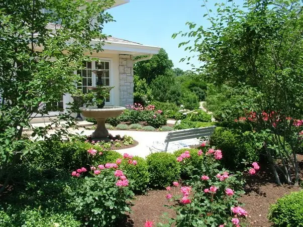 A formal Cincinnati garden by Perrino Landscape with a tiered stone fountain, rose beds and a bench set against a stone home